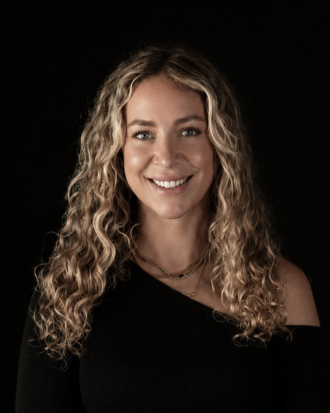 A smiling woman with long curly blonde hair sits against a black background, wearing a black off-the-shoulder long-sleeve top, sheer black tights, and black boots.