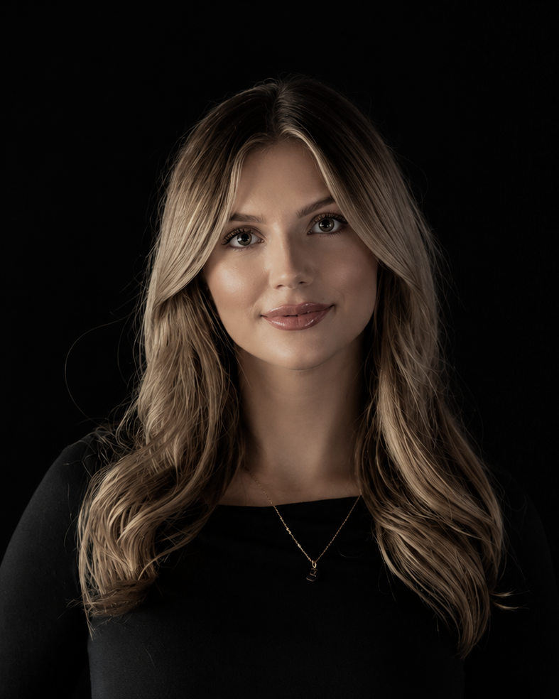 A woman with long, wavy light brown hair with blonde highlights, wearing a black top and a delicate gold necklace, set against a solid black background.