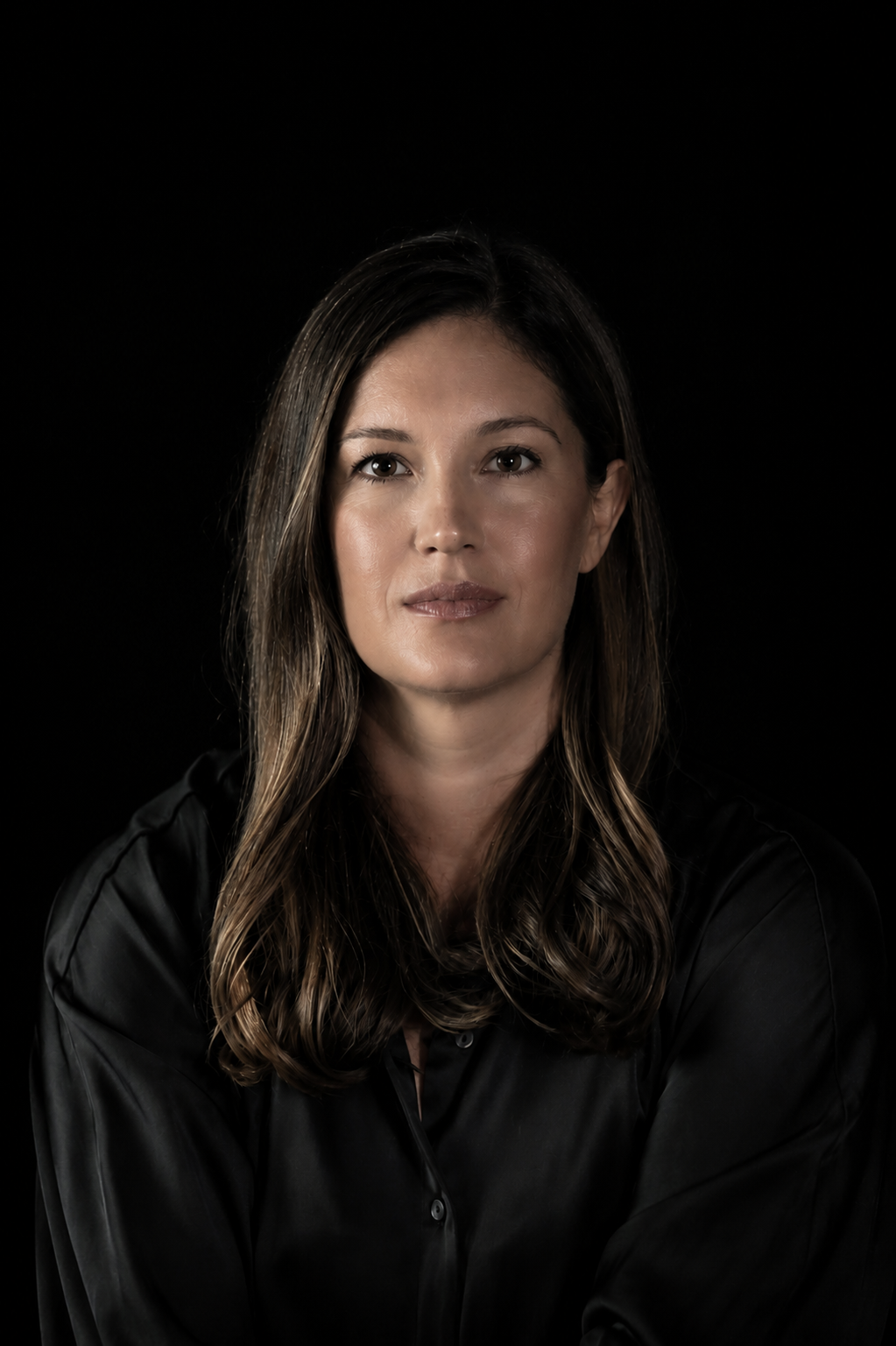 A woman with long dark brown hair wearing a black satin button-up shirt, photographed against a solid black background.