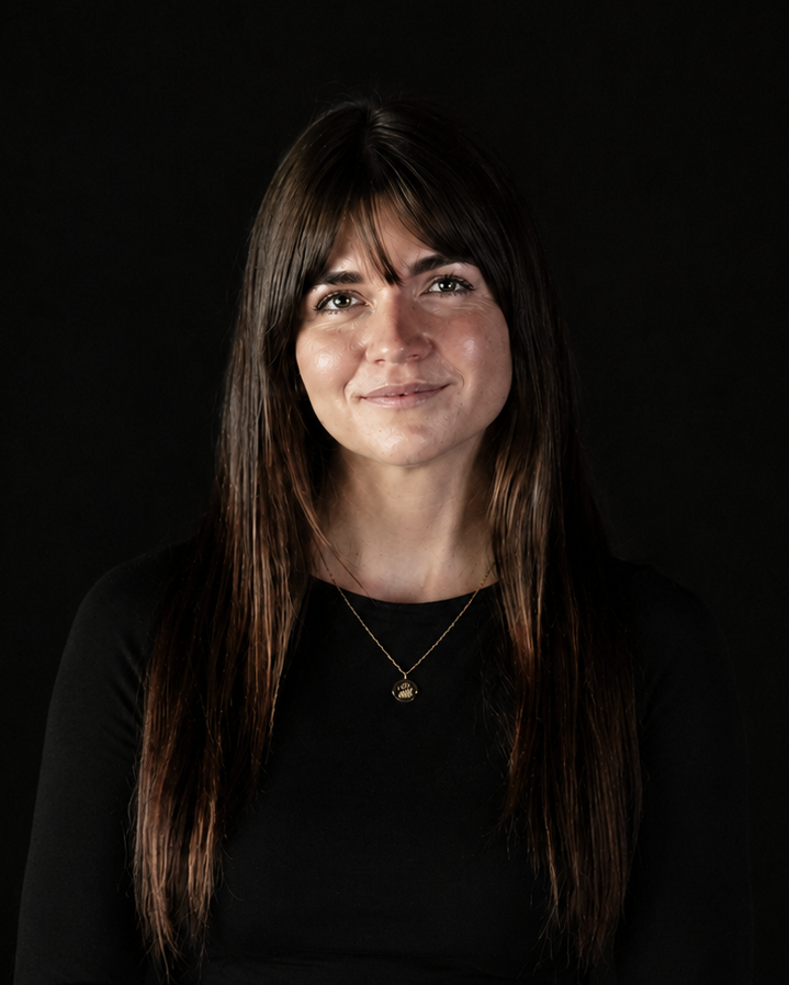 A woman with long dark brown hair sits against a black background, wearing a black long-sleeve top and black trousers, accessorized with a delicate gold necklace.