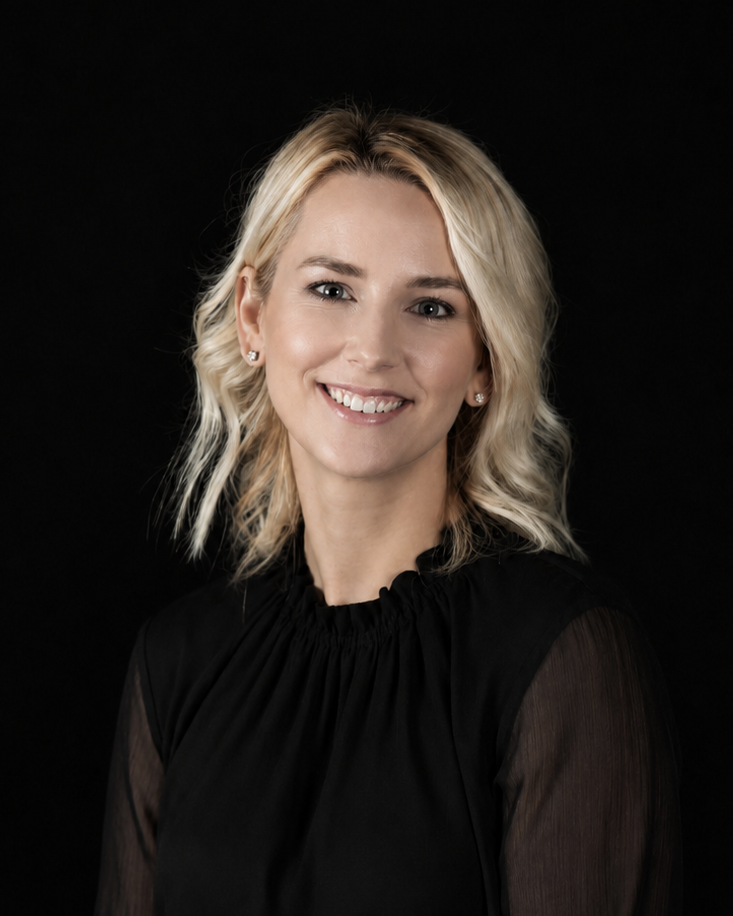 A smiling blonde woman wearing a black sheer long-sleeve blouse, photographed against a solid black background.
