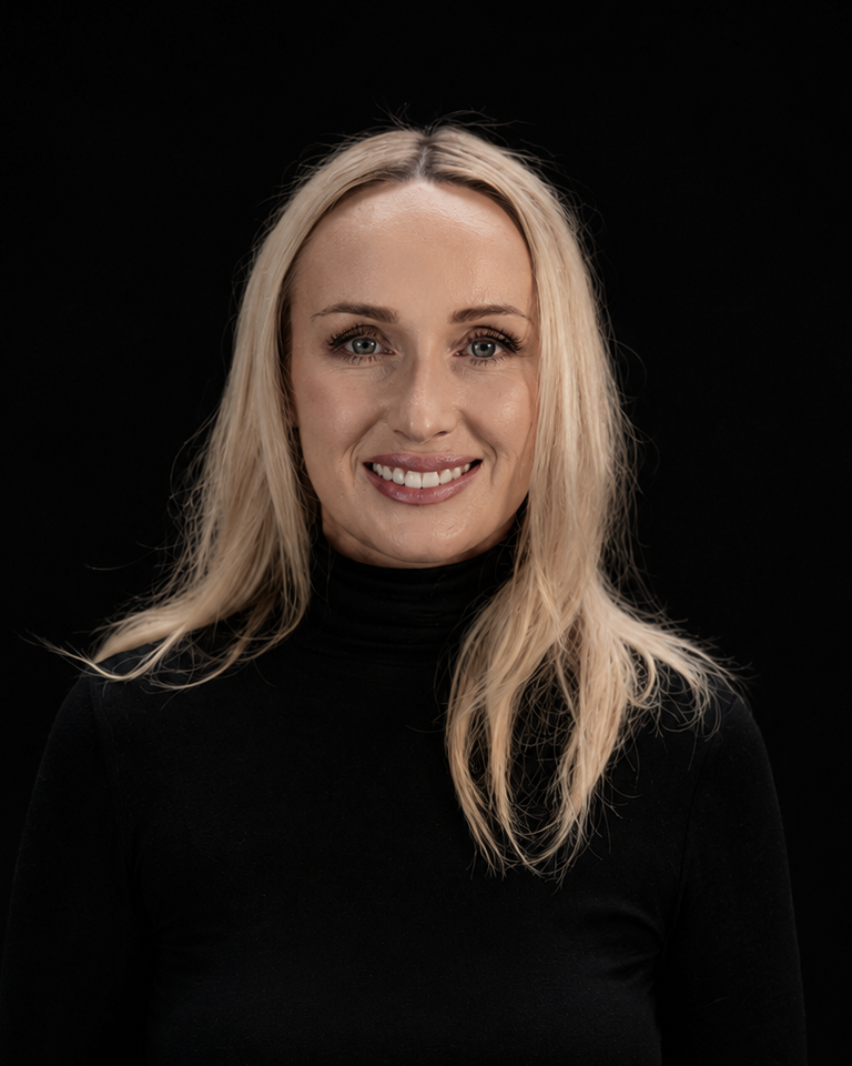 A smiling blonde woman with blue eyes wearing a black turtleneck sweater, photographed against a solid black background.