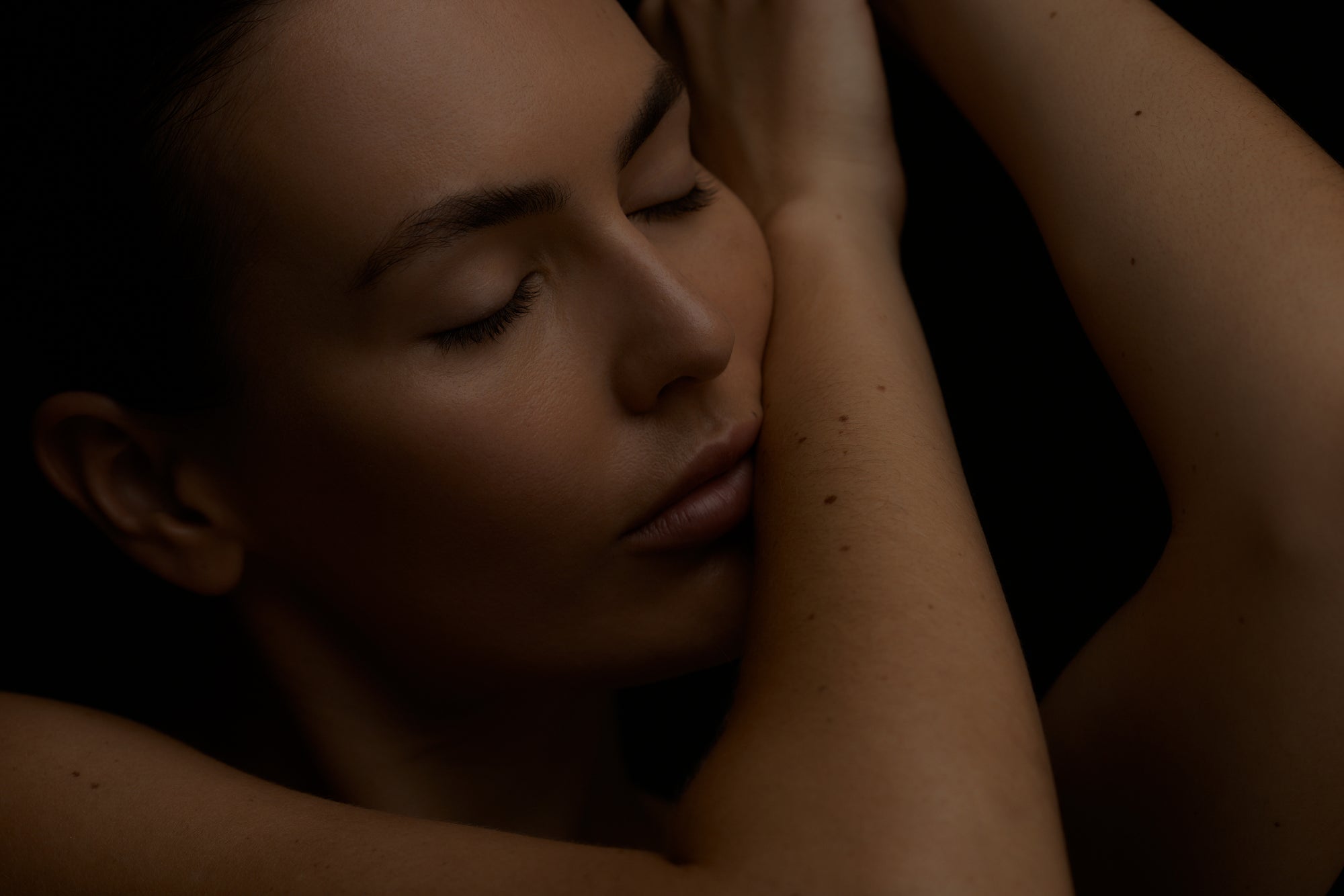 Woman with eyes closed, embracing herself with one arm, against a dark background