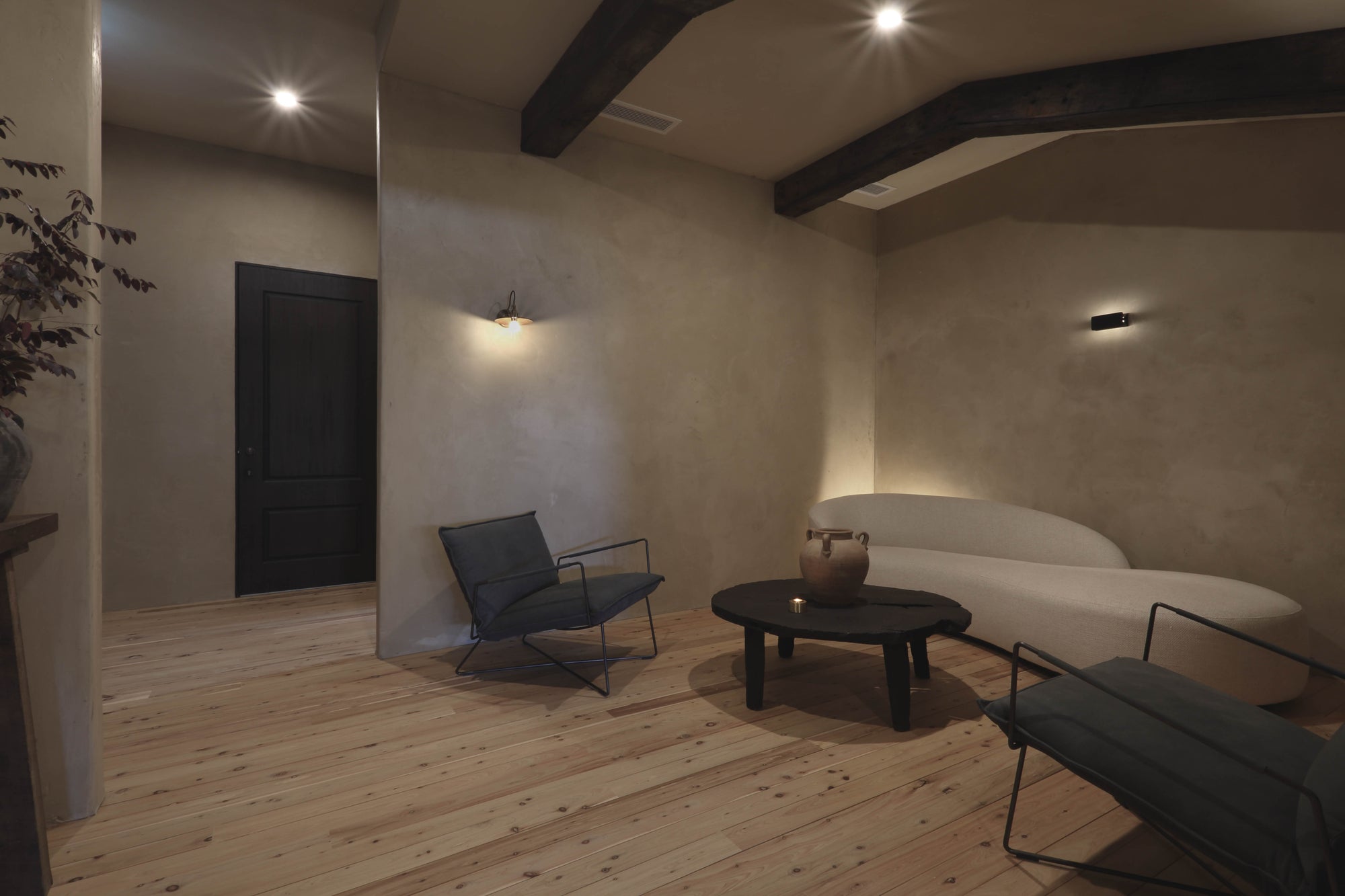 A modern clinic waiting room with light wood floors, textured beige walls, dark ceiling beams, a curved white sofa, a black round coffee table, and two dark gray lounge chairs.
