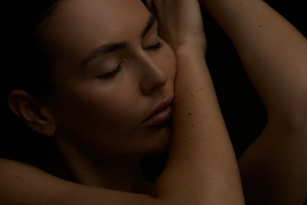 Woman with eyes closed, embracing herself with one arm, against a dark background