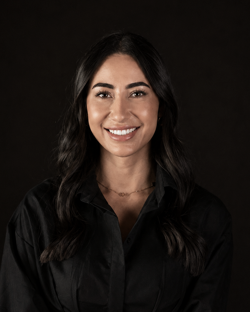A smiling woman with long dark hair wearing a black collared shirt dress with a belt, photographed against a solid black background.